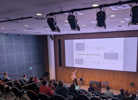 Speaker presenting data charts on a large screen to an audience in a lecture hall.